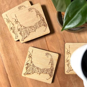 May include: Three square light-colored wooden coasters featuring a laser-engraved map of Cape Cod, Massachusetts. Each coaster displays detailed town names, historical dates, and a compass rose pointing north, rendered in dark brown lines.