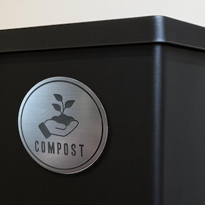 May include: A matte black metal bin featuring a circular silver-toned label. The label displays a graphic of a hand holding a small plant with three leaves and the word COMPOST printed in a bold, sans-serif font below the illustration.