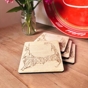May include: A stack of square light-colored wooden coasters featuring a laser-engraved map of Cape Cod, Massachusetts. Each coaster displays detailed geographical outlines, town names, and historical dates etched into the wood surface.