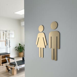 K&ouml;nnte beinhalten: Zwei geb&uuml;rstete goldfarbene Metallschilder f&uuml;r Toiletten an einer hellgrauen Wand. Das linke Schild zeigt eine stilisierte Figur mit Kleid, das rechte Schild eine stilisierte Figur mit geraden Beinen als klassische Symbole.