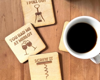 May include: Three square light-colored wooden coasters with dark engraved text and graphics. One reads I PULL OUT with a corkscrew, one says YOU HAD ME AT MERLOT with two wine glasses, and one says SCREW IT with a corkscrew graphic.