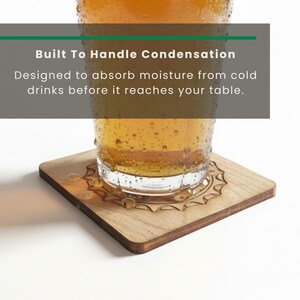 May include: A square wooden coaster with a sunburst engraving, supporting a glass of amber liquid covered in condensation. Text overlay reads: Built To Handle Condensation. Designed to absorb moisture from cold drinks before it reaches your table.