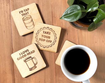 May include: Four square light-colored wooden coasters with dark brown laser-engraved designs. Three coasters feature beer-related illustrations and text: I'D TAP THAT, TAKE YOUR TOP OFF, and I LOVE GOOD HEAD. One coaster is blank and partially covered.