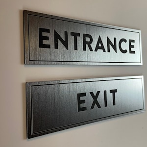May include: Two rectangular signs with a brushed metallic finish and black borders. The top sign features the word ENTRANCE in bold black capital letters, while the bottom sign displays the word EXIT in the same bold black capital lettering style.