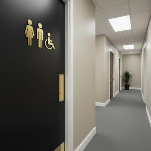 K&ouml;nnte beinhalten: Eine schwarze T&uuml;r mit goldfarbenen Schildern, die eine Frau, einen Mann und eine Person im Rollstuhl darstellen. Die T&uuml;r verf&uuml;gt &uuml;ber eine goldfarbene Trittplatte und einen vertikalen T&uuml;rgriff in einem Flur mit beigen W&auml;nden.