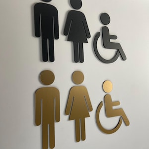 May include: A set of six wall-mounted restroom signs arranged in two rows. The top row features black icons for a person, a person in a dress, and a wheelchair user. The bottom row displays identical icons in a metallic gold finish.