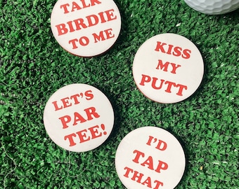 Può includere: Quattro sottobicchieri circolari bianchi con testo rosso. Il testo recita: TALK BIRDIE TO ME, KISS MY PUTT, LET'S PAR TEE! e I'D TAP THAT. Ogni sottobicchiere presenta una superficie liscia e un bordo rotondo, presentati come un set di accessori.