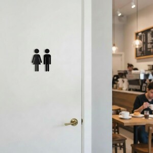 K&ouml;nnte beinhalten: Eine wei&szlig;e T&uuml;r mit einem schwarzen Toilettenschild, das stilisierte Symbole einer Person in einem Kleid und einer Person in einer Hose zeigt, angebracht &uuml;ber einem messingfarbenen T&uuml;rgriff mit runder Rosette.