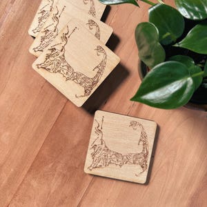 May include: A set of square light-colored wooden coasters, each featuring a dark brown laser-engraved map of Cape Cod, Massachusetts. The map includes detailed geographic outlines, town names, and historical dates etched into the wood surface.