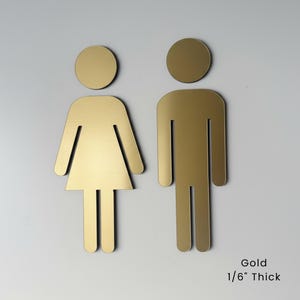 K&ouml;nnte beinhalten: Zwei goldfarbene Toilettenschilder mit stilisierten menschlichen Figuren, eine mit dreieckiger Rockform und eine mit rechteckiger K&ouml;rperform. Jedes Schild hat einen runden Kopf und ist laut Text 1/6 Zoll dick.