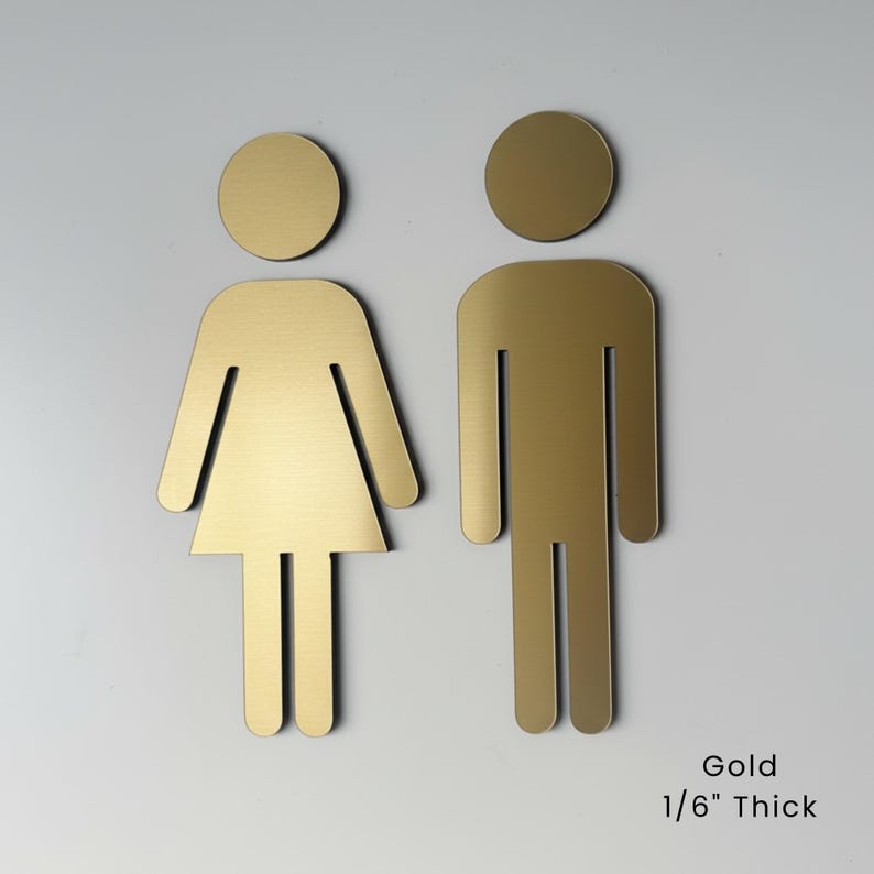 K&ouml;nnte beinhalten: Zwei goldfarbene Toilettenschilder mit stilisierten menschlichen Figuren, eine mit dreieckiger Rockform und eine mit rechteckiger K&ouml;rperform. Jedes Schild hat einen runden Kopf und ist laut Text 1/6 Zoll dick.