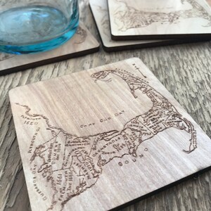 May include: A square wooden coaster featuring a laser-engraved map of Cape Cod, Massachusetts. The map includes detailed labels for towns like Plymouth, Barnstable, and Falmouth, along with dates and geographical features like Cape Cod Bay.