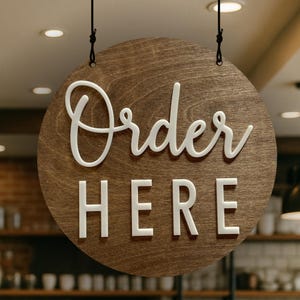 May include: A circular wooden sign with a dark brown wood grain finish, suspended by two black cords. The sign features the white text "Order" in a cursive script font above the white text "HERE" in a bold, sans-serif block font.