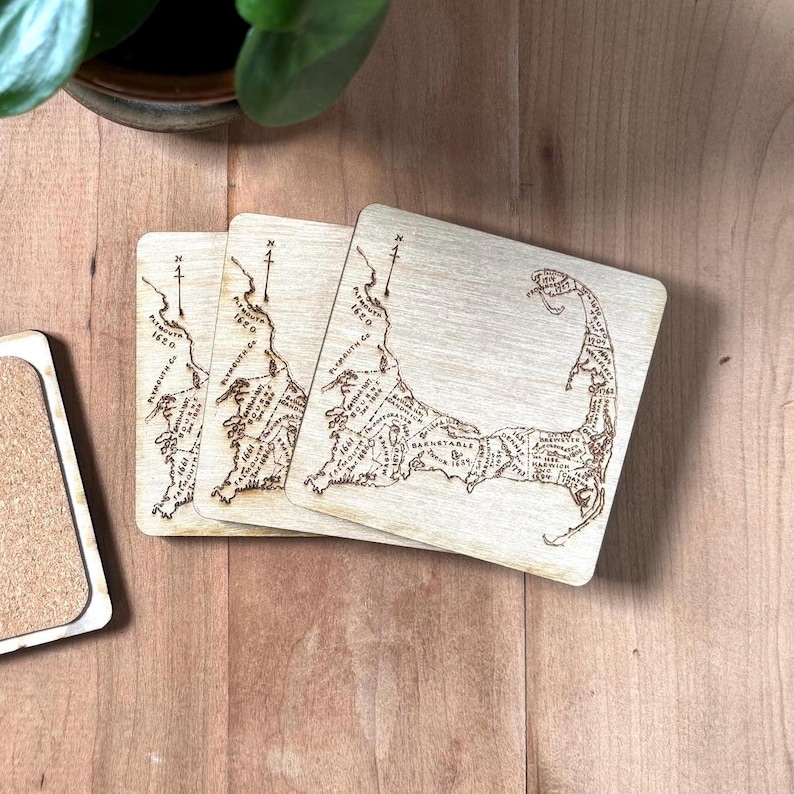 May include: Three square light-colored wooden coasters featuring a laser-engraved map of Cape Cod, Massachusetts. Each coaster displays detailed geographical outlines, town names, and historical dates, with a compass rose pointing north.