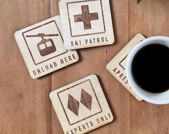 May include: Four square wooden coasters featuring dark brown laser-engraved ski resort signage. Designs include a ski patrol cross, a gondola with the text UNLOAD HERE, two diamonds with EXPERTS ONLY, and a partial coaster with the text APRES.