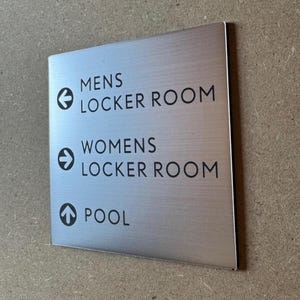 May include: A silver metal sign with black text and arrows indicating directions to the mens and womens locker rooms and the pool. The sign reads "Mens Locker Room", "Womens Locker Room", and "Pool".