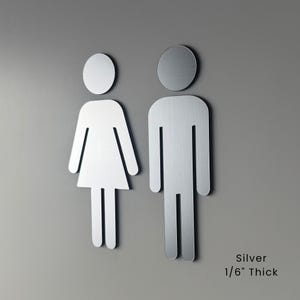 K&ouml;nnte beinhalten: Zwei silberne Toilettenschilder mit stilisierten menschlichen Figuren, eine mit Kleiderform und eine mit rechteckiger Form. Die Schilder haben eine geb&uuml;rstete Metalloberfl&auml;che und sind 0,42 cm dick, wie auf der Oberfl&auml;che angegeben.