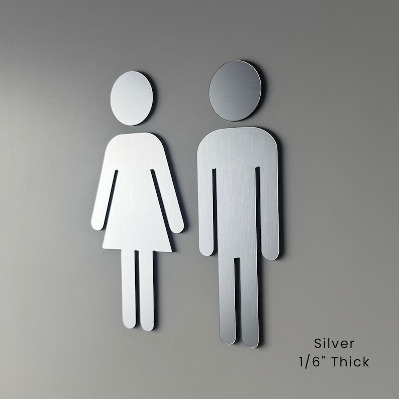 K&ouml;nnte beinhalten: Zwei silberne Toilettenschilder mit stilisierten menschlichen Figuren, eine mit Kleiderform und eine mit rechteckiger Form. Die Schilder haben eine geb&uuml;rstete Metalloberfl&auml;che und sind 0,42 cm dick, wie auf der Oberfl&auml;che angegeben.
