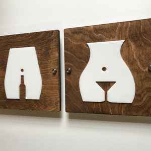 Könnte beinhalten: Zwei quadratische Wandtafeln aus Holz mit weißen Acrylausschnitten in Form menschlicher Torsi. Die linke Tafel zeigt die Silhouette einer Bierflasche, während die rechte Tafel die Silhouette eines Martiniglases darstellt, befestigt mit Metall.