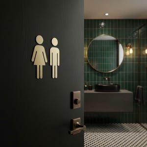 K&ouml;nnte beinhalten: Eine mattschwarze T&uuml;r mit goldenen, metallischen Toilettensymbolen einer Person im Kleid und einer Person in einer Hose. Die T&uuml;r verf&uuml;gt &uuml;ber ein passendes rechteckiges goldenes Schlossblech und einen horizontalen T&uuml;rdr&uuml;cker.