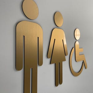 K&ouml;nnte beinhalten: Ein Set aus drei metallisch goldenen Toilettenschildern, bestehend aus einer stilisierten Figur in rechteckiger Form, einer Figur in Kleiderform und einer Figur im Rollstuhl. Die Schilder haben eine glatte, geb&uuml;rstete Oberfl&auml;che.