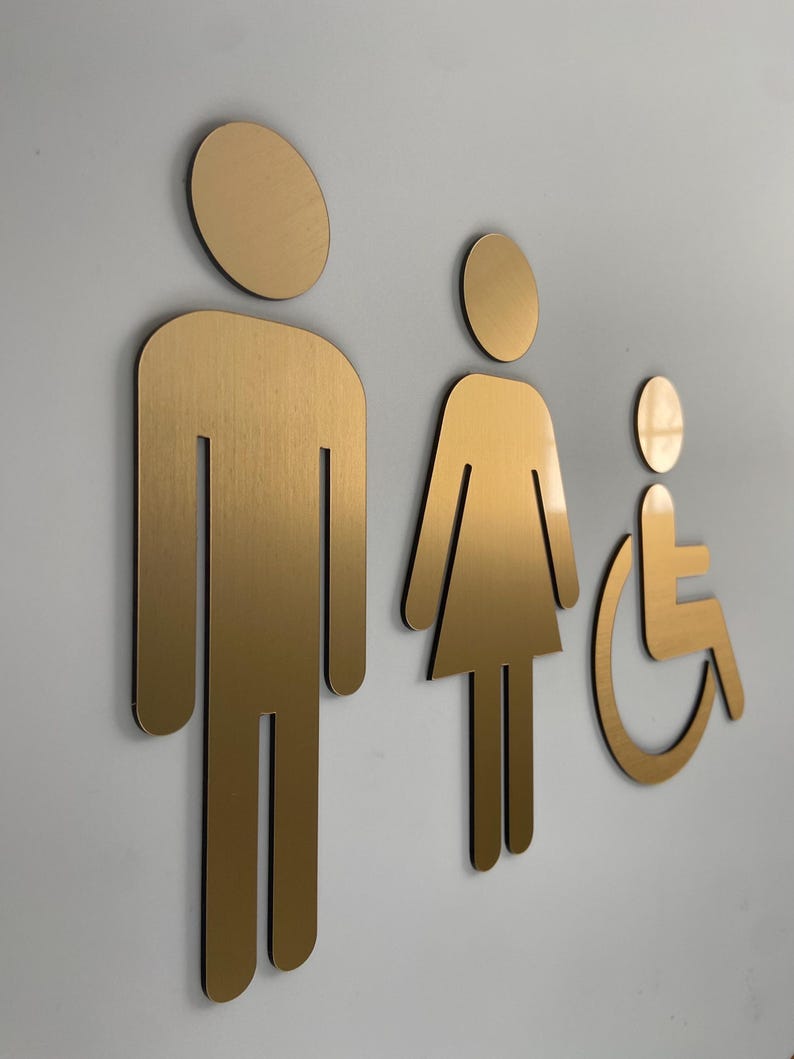 K&ouml;nnte beinhalten: Ein Set aus drei metallisch goldenen Toilettenschildern, bestehend aus einer stilisierten Figur in rechteckiger Form, einer Figur in Kleiderform und einer Figur im Rollstuhl. Die Schilder haben eine glatte, geb&uuml;rstete Oberfl&auml;che.