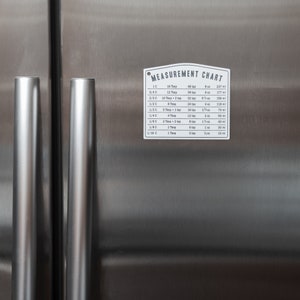 Puede incluir: Un gr&aacute;fico de metal blanco con el texto "MEASUREMENT CHART" impreso en negro. El gr&aacute;fico enumera las medidas equivalentes para tazas y cucharadas en onzas y mililitros.