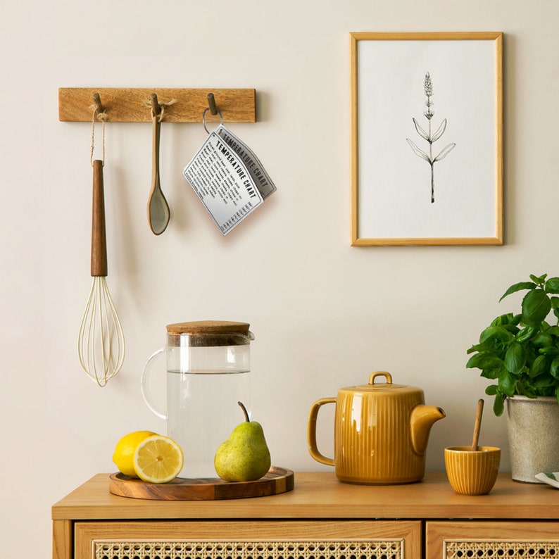 Puede incluir: Un mostrador de cocina con una tabla de cortar de madera, una jarra de vidrio con agua, una tetera amarilla, un taz&oacute;n amarillo peque&ntilde;o, una pera, dos limones y una impresi&oacute;n enmarcada de una planta. El mostrador tiene un estante de madera con un gancho de metal que sostiene una tarjeta de metal con el texto "TEMPERATURE CHART" impreso en ella.