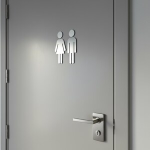 K&ouml;nnte beinhalten: Eine hellgraue T&uuml;r mit einem metallisch silbernen Toilettenschild, das zwei stilisierte Figuren zeigt, eine mit Kleid und eine mit Hose. Die T&uuml;r ist mit einem passenden silbernen T&uuml;rdr&uuml;cker mit Schl&uuml;sselloch und drei Scharnieren versehen.