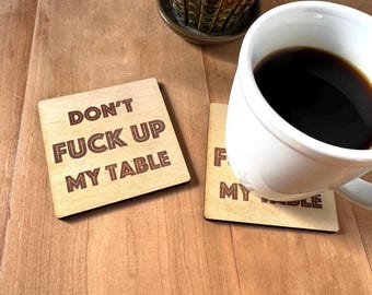 May include: Two square light-colored wooden coasters with dark brown engraved text that reads "DON'T FUCK UP MY TABLE". One coaster is placed flat, while the other is partially covered by a white ceramic mug filled with black coffee.