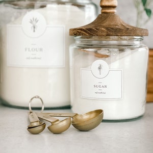 May include: Two glass canisters with wooden lids, one labeled "Flour" and the other "Sugar", both with a simple plant illustration. The canisters are filled with white powder and are sitting on a white countertop. Three gold measuring spoons are in the foreground.