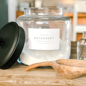 May include: A clear glass jar with a black lid filled with white laundry detergent. The jar has a white label that reads "Laundry Detergent Clean & Deodorize". A wooden scoop and a clear glass vase with a sprig of greenery are also in the image.