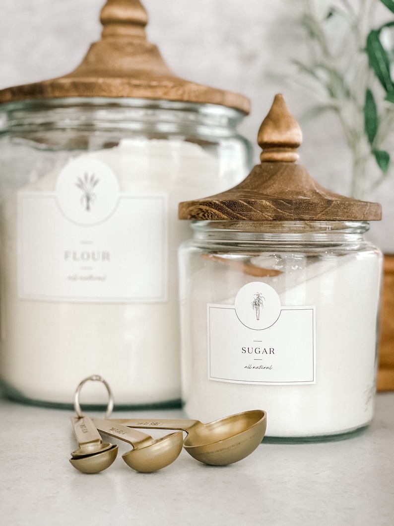 May include: Two glass canisters with wooden lids, one labeled "Flour" and the other "Sugar", both with a simple plant illustration. The canisters are filled with white powder and are sitting on a white countertop. Three gold measuring spoons are in the foreground.