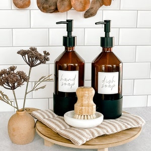 May include: Two amber glass pump bottles with black pumps and labels that read "hand soap" and "dish soap". The bottles are on a wooden tray with a brown and white striped fabric pad and a white ceramic dish with a wooden brush.