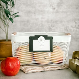 May include: A white wire basket with a black label that says "APPLES - eat natural" and a wooden handle. The basket is filled with red apples and is sitting on a brown and white striped mat.
