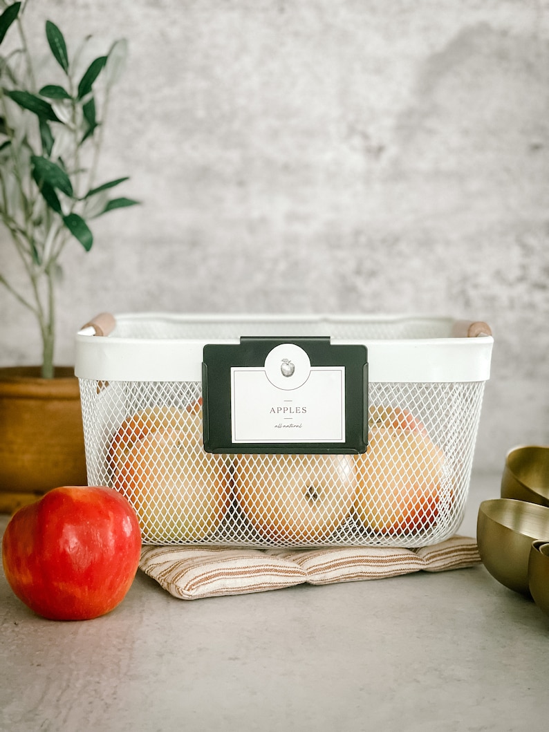 May include: A white wire basket with a black label that says "APPLES - eat natural" and a wooden handle. The basket is filled with red apples and is sitting on a brown and white striped mat.