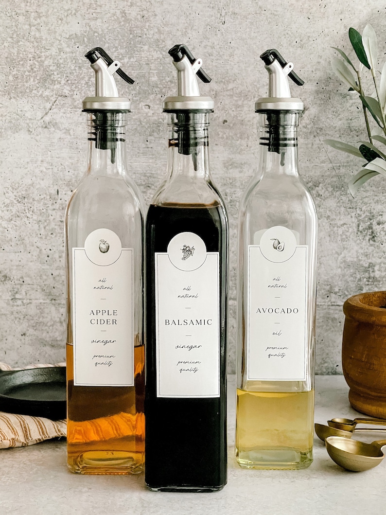 Puede incluir: Tres botellas de vidrio con etiquetas y boquillas de vertido. Las botellas est&aacute;n llenas de diferentes l&iacute;quidos: vinagre de sidra de manzana, vinagre bals&aacute;mico y aceite de aguacate. Las etiquetas son blancas con texto negro e ilustraciones simples de una manzana, una uva y un aguacate.
