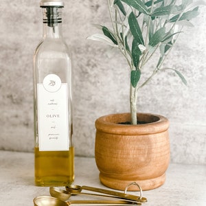 Puede incluir: Una botella de vidrio con un pico vertedor negro llena de aceite de oliva. La botella tiene una etiqueta que dice "all natural OLIVE oil premium quality". La botella est&aacute; al lado de un peque&ntilde;o olivo en una maceta de madera y tres cucharaditas doradas.