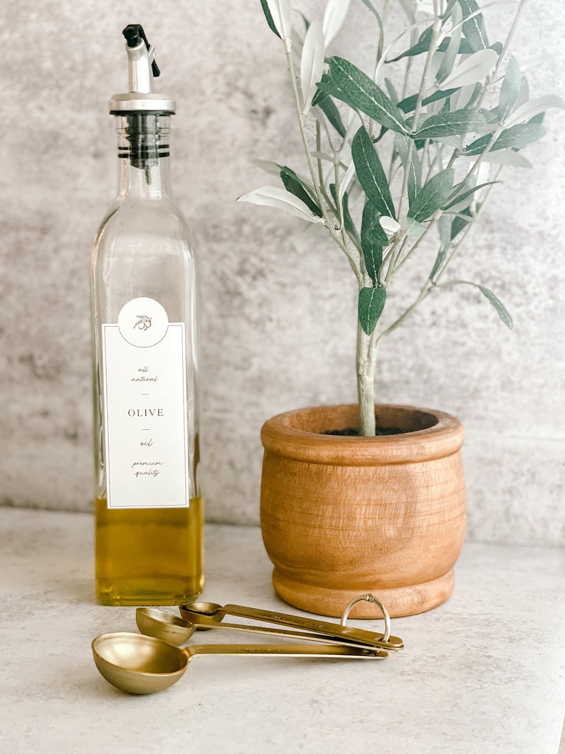 Puede incluir: Una botella de vidrio con un pico vertedor negro llena de aceite de oliva. La botella tiene una etiqueta que dice "all natural OLIVE oil premium quality". La botella est&aacute; al lado de un peque&ntilde;o olivo en una maceta de madera y tres cucharaditas doradas.