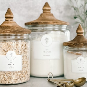 May include: Three glass canisters with wooden lids and labels. The canisters are filled with oats, flour, and sugar. The labels have a simple design with the name of the ingredient and a small graphic of a plant.