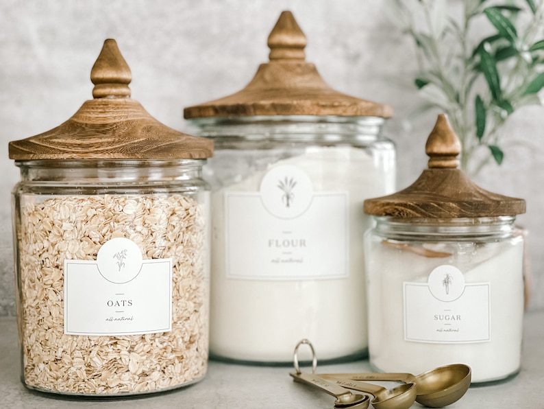 May include: Three glass canisters with wooden lids and labels. The canisters are filled with oats, flour, and sugar. The labels have a simple design with the name of the ingredient and a small graphic of a plant.