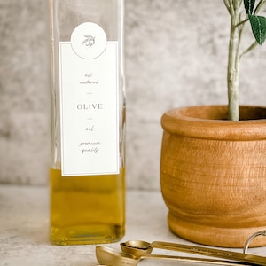 Puede incluir: Una botella de vidrio transparente llena de aceite de oliva dorado. La botella tiene una etiqueta blanca con el texto "all natural - OLIVE oil - premium quality". La botella est&aacute; sobre una superficie gris claro junto a un peque&ntilde;o olivo en maceta y tres cucharas medidoras doradas.