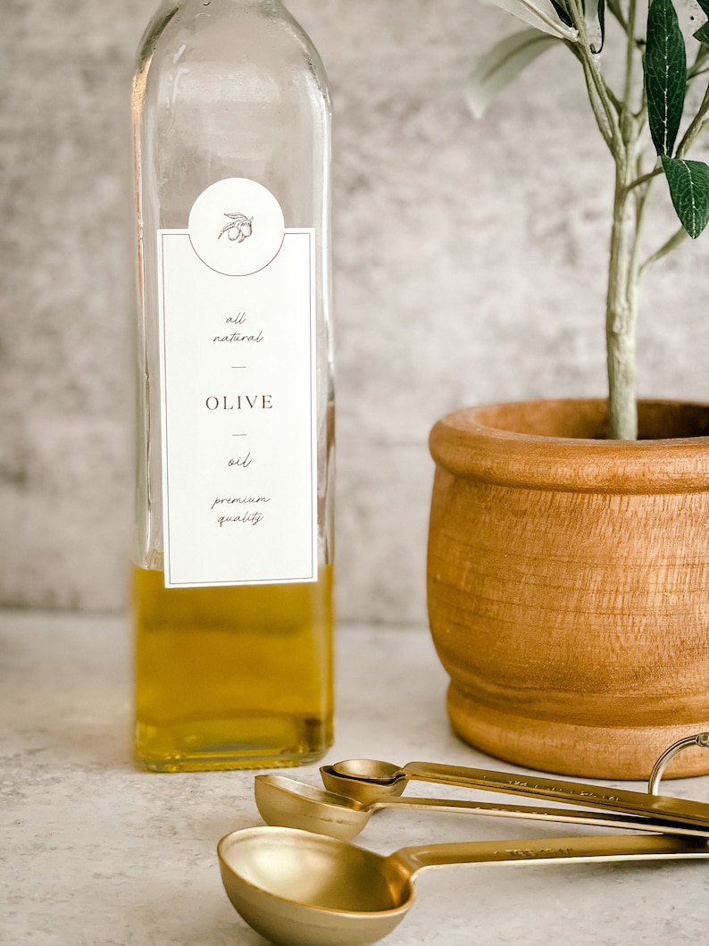 Puede incluir: Una botella de vidrio transparente llena de aceite de oliva dorado. La botella tiene una etiqueta blanca con el texto "all natural - OLIVE oil - premium quality". La botella est&aacute; sobre una superficie gris claro junto a un peque&ntilde;o olivo en maceta y tres cucharas medidoras doradas.