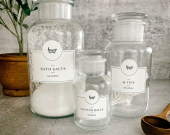Può includere: Tre barattoli di vetro trasparente con etichette bianche e testo nero. Le etichette riportano "Bath Salts", "Cotton Balls" e "Q-Tips". Ogni etichetta presenta una piccola illustrazione di una vasca da bagno. Tutte le etichette includono il testo "all natural".