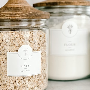 May include: Two glass jars with wooden lids, one filled with oats and labeled "OATS all natural" and the other filled with flour and labeled "FLOUR all natural".
