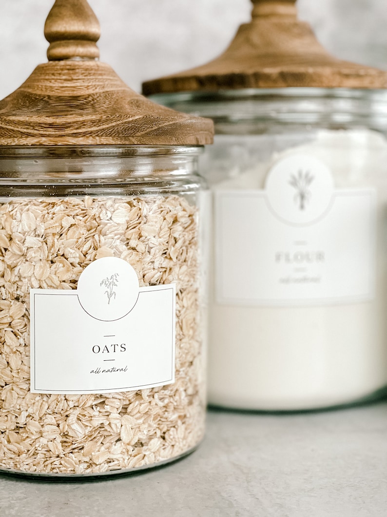 May include: Two glass jars with wooden lids, one filled with oats and labeled "OATS all natural" and the other filled with flour and labeled "FLOUR all natural".