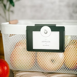 May include: White wire basket with a black label that says "Apples - all natural" with a small apple graphic. The basket is filled with red apples.