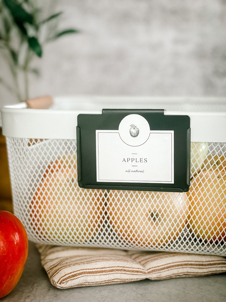 May include: White wire basket with a black label that says "Apples - all natural" with a small apple graphic. The basket is filled with red apples.