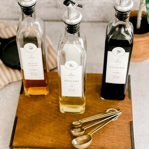 Puede incluir: Tres botellas de vidrio con boquillas y etiquetas en una bandeja de madera. Las etiquetas dicen "Vinagre de sidra de manzana", "Aceite de aguacate" y "Vinagre bals&aacute;mico". La bandeja tiene la palabra "ACEITE" grabada. Cuatro cucharas medidoras doradas est&aacute;n en la bandeja.