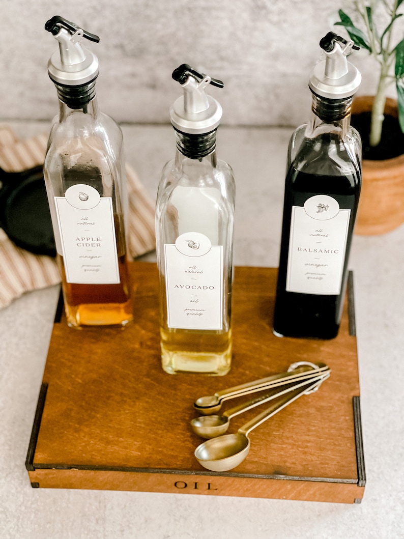 Puede incluir: Tres botellas de vidrio con boquillas y etiquetas en una bandeja de madera. Las etiquetas dicen "Vinagre de sidra de manzana", "Aceite de aguacate" y "Vinagre bals&aacute;mico". La bandeja tiene la palabra "ACEITE" grabada. Cuatro cucharas medidoras doradas est&aacute;n en la bandeja.